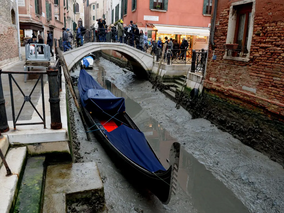 L'image montre un canal dans la ville de Venise, en Italie.  C'est sec et il y a un bateau sur le lit du canal.  en arrière-plan, les gens traversent un petit pont à pied et regardent le canal vide. 