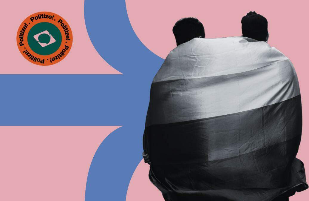 A imagem mostra duas pessoas cobertas com uma grande bandeira LGBTI+